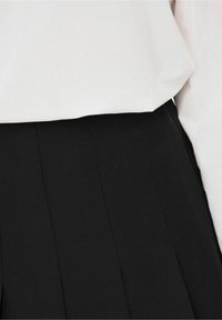 Gros plan d'une personne portant un haut blanc rentré dans un pantalon noir taille haute avec des détails de coutures verticales.