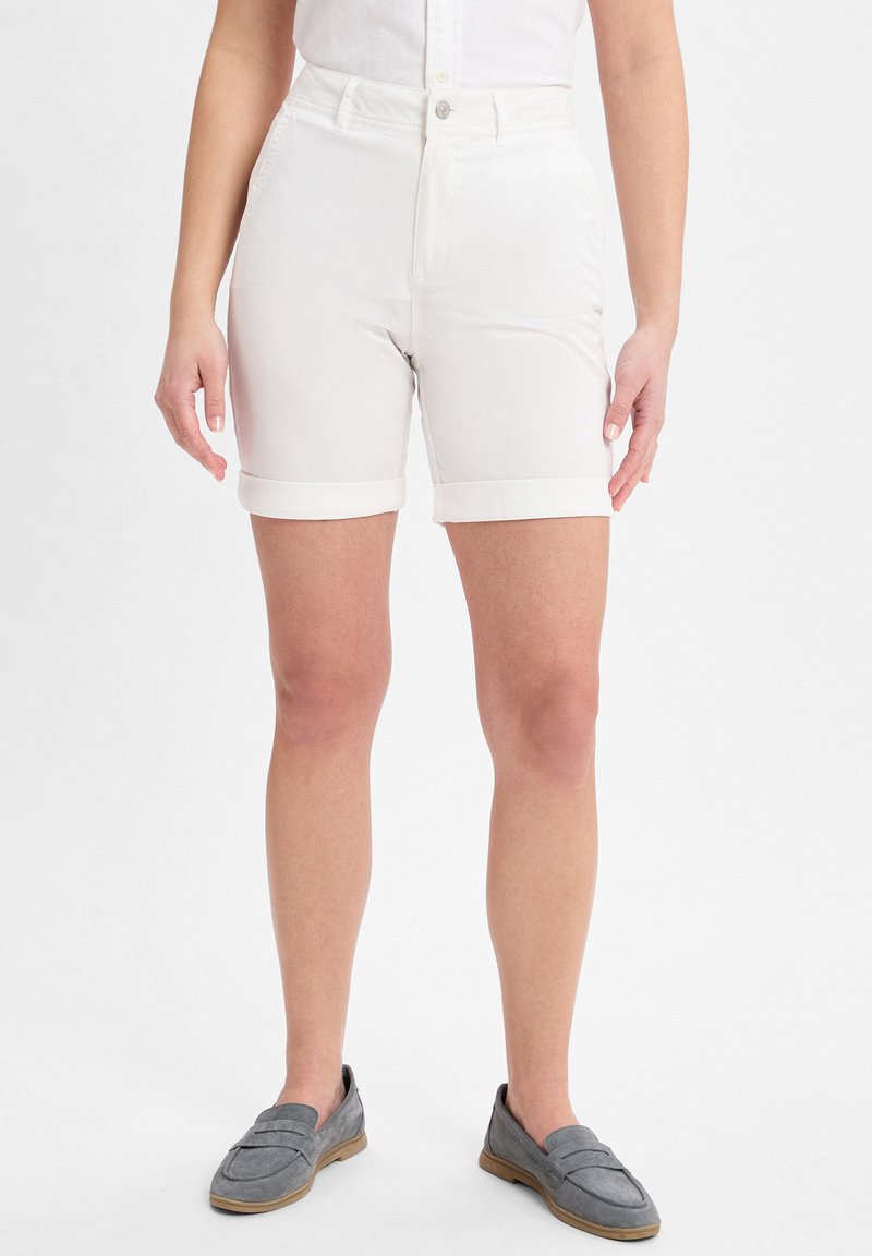 Weiße Baumwollshorts mit hoher Taille, aufgerollten Säumen und vorderen Taschen. Kombiniert mit grauen Wildledermokassins. Einfarbiger Hintergrund.