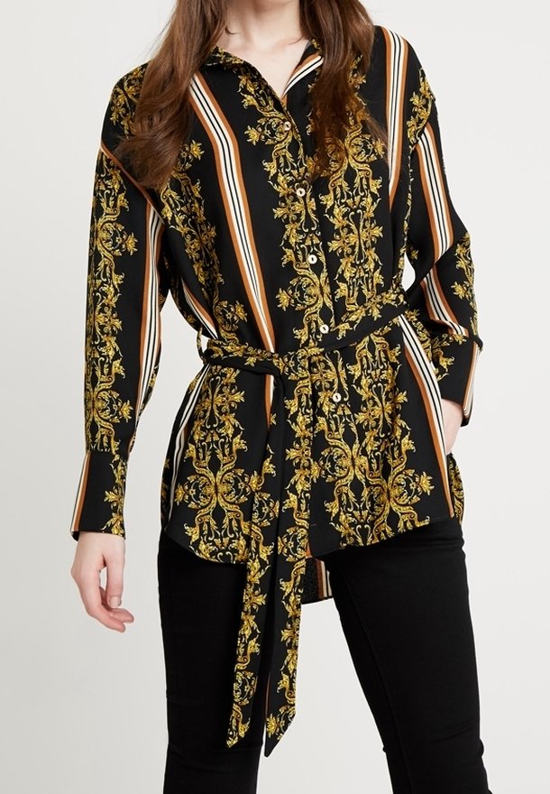 Femme portant une chemise noire boutonnée avec des motifs floraux baroques dorés, des rayures verticales beige et blanches, une ceinture en tissu et un pantalon noir.
