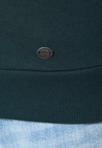 Tmavě zelená mikina z materiálu s žebrovým lemem a malým oválným kovovým logem. Pozadí ukazuje světle modrou texturu denimu.