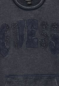 Tumedast hall sweatpusa, millel on tekstuuriga "GUESS" kiri, ribidega ümar kaelus ja siledapinnaline eesmine tasku.
