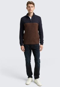 Junger Mann, der einen navy- und braunfarbenen Half-Zip-Pullover, dunkle Jeans und weiße Sneakers vor einem schlichten Hintergrund trägt.