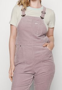 Rosa kordflöjelsoveraller med justerbara axelremmar, framficka och sidofickor. Bärs över en randig skjorta i pastellfärger.