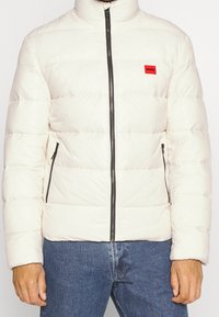 Hellbeige Daunenjacke mit hohem Kragen, horizontalen Steppnähten, schwarzen Reißverschlüssen und einem kleinen roten Logo auf der Brust. Normale Passform.