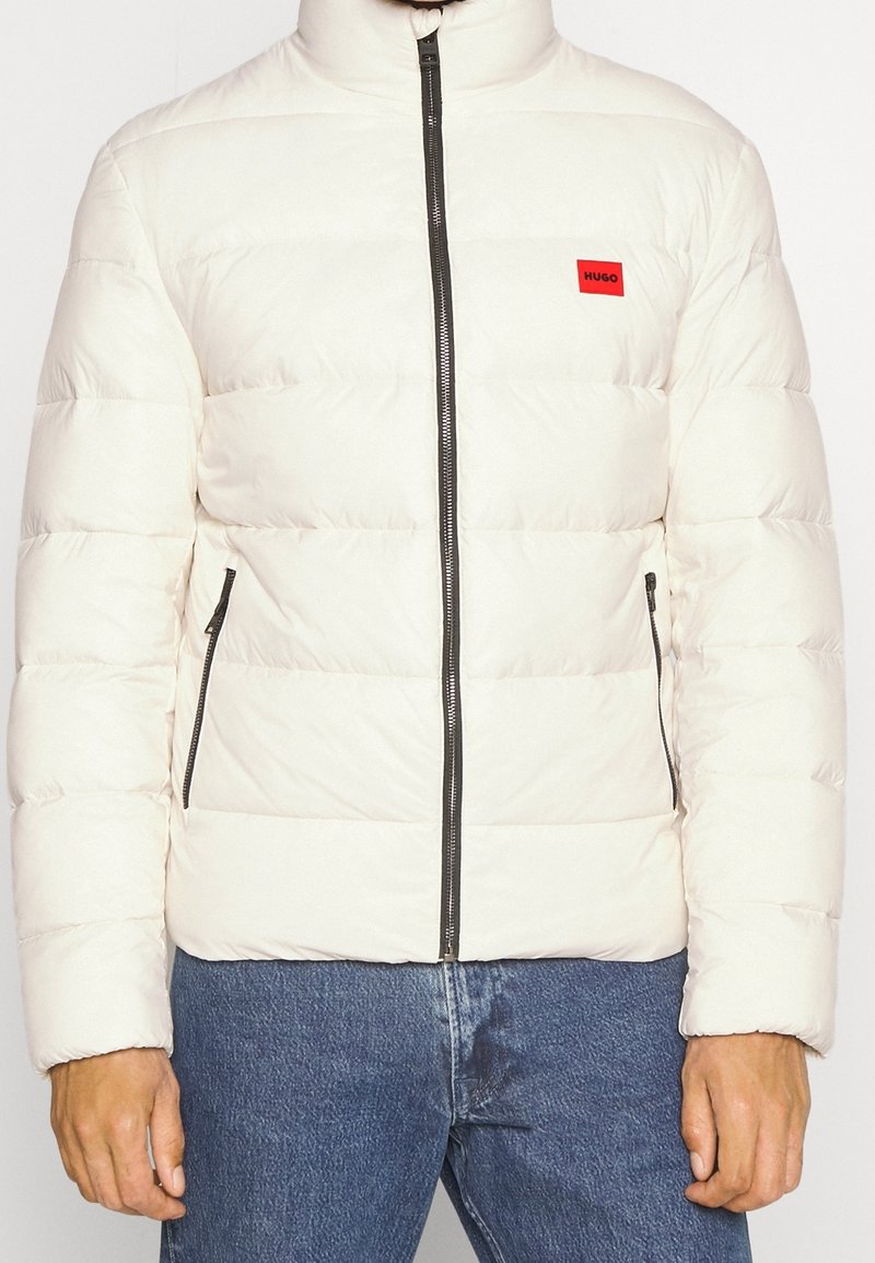 Hellbeige Daunenjacke mit hohem Kragen, horizontalen Steppnähten, schwarzen Reißverschlüssen und einem kleinen roten Logo auf der Brust. Normale Passform.