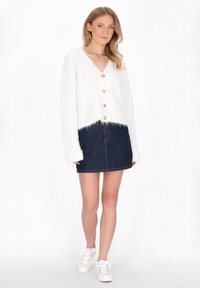 Bílý pletený cardigan s výstřihem do V, zdobený pěti zlatými knoflíky, nošený přes tmavé džíny a bílé tenisky. Měkká textura, zkrácený střih.