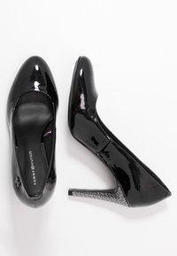 Zapatos de tacón alto de charol negro con punta afilada y tacón plateado texturizado. Logo en la plantilla, diseño elegante y acabado brillante.