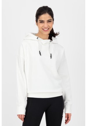 Junge Frau lächelt, trägt einen weißen Kapuzenpullover mit Kordeln und schwarze Leggings, steht vor einem schlichten hellen Hintergrund.