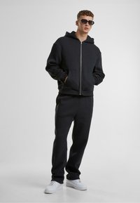 Haut à zip noir avec une poche avant, associé à un pantalon de jogging noir assorti. Porté avec des baskets blanches et des lunettes de soleil noires.