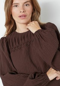 Blusa marrone a pieghe con alto collo e tessuto texturizzato, con maniche arricciate e cuciture decorative sul petto.