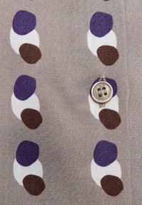 Beige tyg med stora, släta oregelbundna prickar i lila, brunt och vitt; har en rund, ljus knapp.