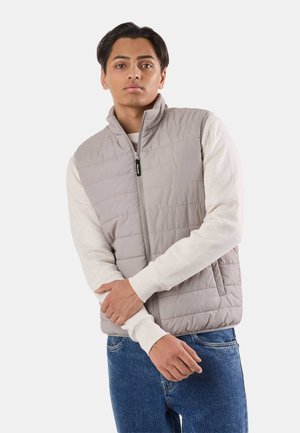 Młody mężczyzna o średnim odcieniu skóry, ubrany w jasnoszarą pikowaną kamizelkę na kremowej koszuli z długim rękawem i niebieskich dżinsach, stoi z założonymi ramionami.