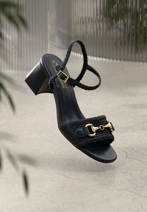 Sort hæleleje sandal med flettede læderremme og detalje med guldspænde, hængende mod en glat grå baggrund med bløde skygger.