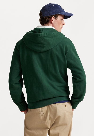 Homem vestindo um moletom verde escuro, calças bege, camisa de colarinho branca e um boné de corduroy azul-marinho, de lado com as mãos nos bolsos.