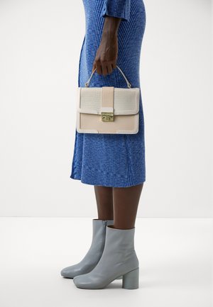 Personne portant une robe côtelée bleue et des bottines grises, tenant un sac à main beige avec un fermoir doré par sa poignée supérieure.