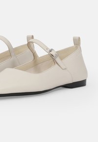 Bailarinas de cuero beige con punta afilada, correa ajustable en el tobillo con hebilla y un bajo tacón negro. Textura suave con un detalle mínimo.