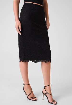 Jupe midi en dentelle noire avec ourlet festonné portée avec des sandales noires à talons hauts et bretelles sur un fond clair.