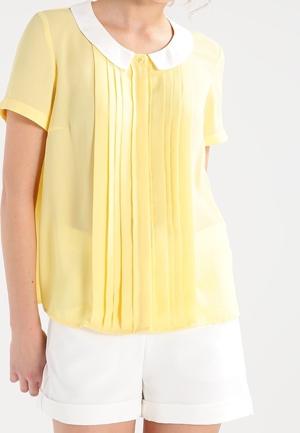 Femme portant un chemisier jaune à manches courtes avec col blanc et plis, associé à un short blanc à ourlet roulé sur un fond uni.