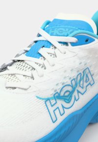 Hvite sneakers med blå detaljer, med mesh-overdel, teksturert såle, og tydelig HOKA-profilering på siden.