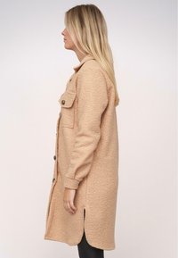 Beige kabát s dlouhým rukávem z texturované tkaniny, na knoflíky, se dvěma náprsními kapsami a bočními rozparky. Přiléhavý střih a délka pod kolena.