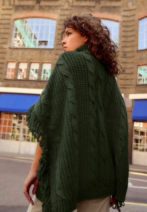 Waldgrün gestrickter Poncho mit Zopfmustern, Fransenrändern und strukturiertem Gewebe, getragen über einem hellen Outfit vor einer städtischen Kulisse.