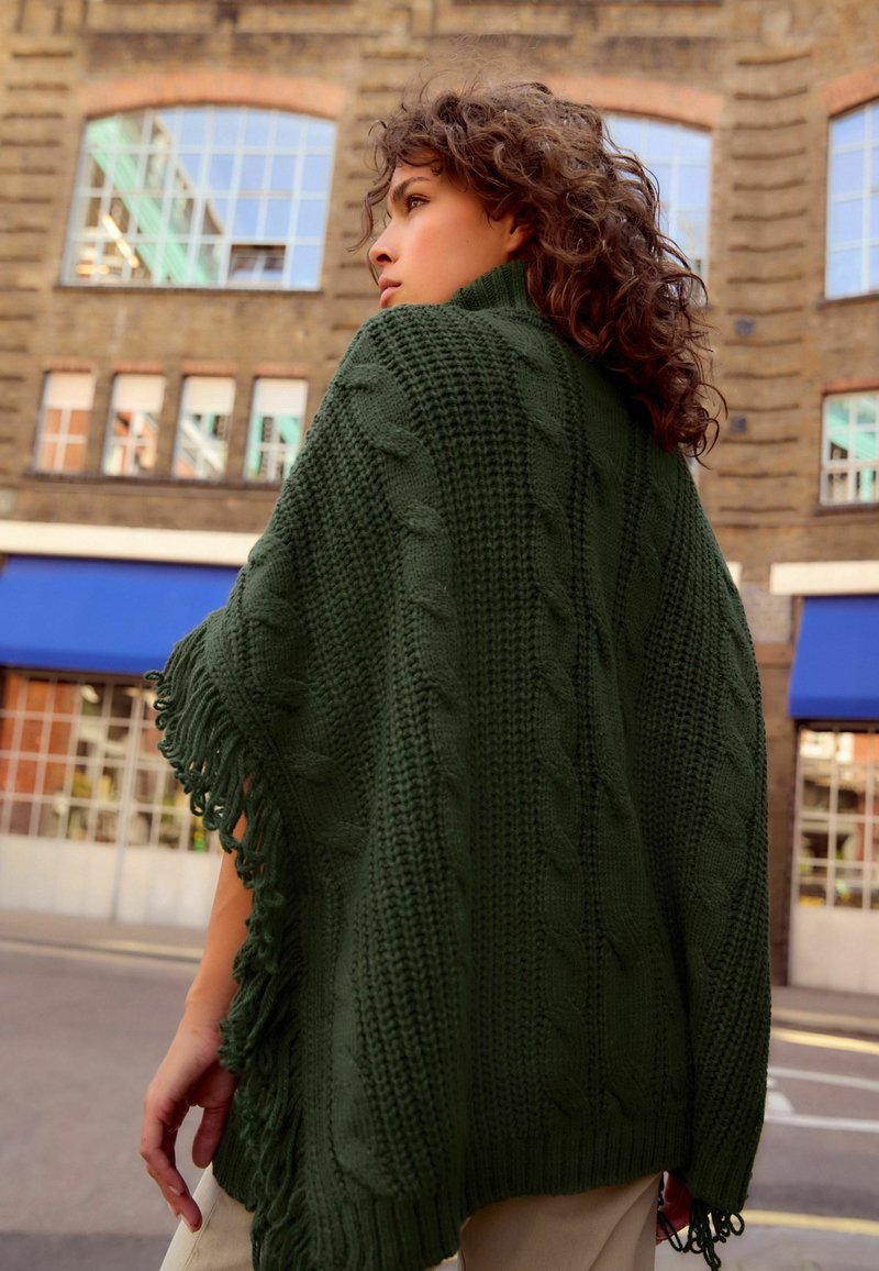 Skovgrøn strikket poncho med kabelmønstre, frynser og struktureret væv, båret over et lyst outfit mod en bybaggrund.