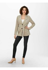 Blazer in corduroy beige con vita regolabile, colletto a revers e due tasche frontali, abbinato a jeans skinny neri e tacchi nudi.