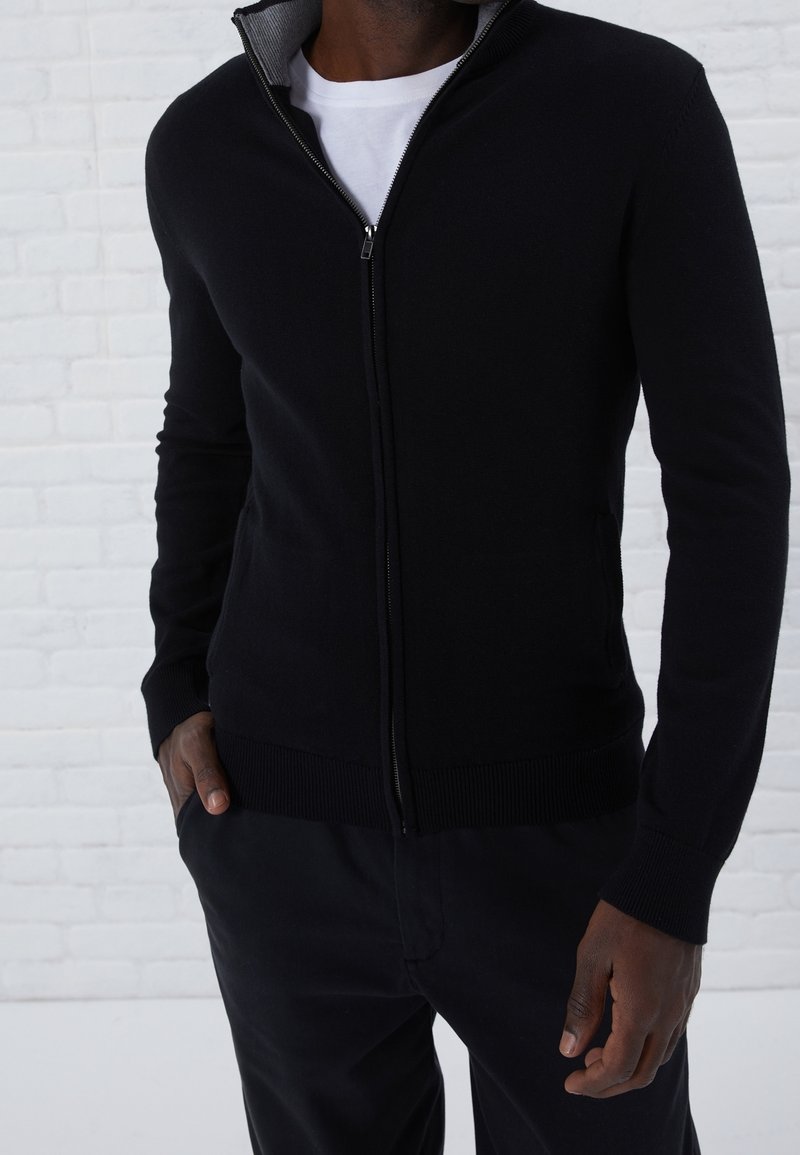 Homme portant un pull noir à fermeture éclair sur un t-shirt blanc, les mains détendues, debout devant un mur de briques blanc.