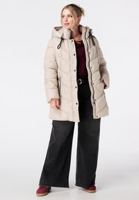 Beige gewatteerde jas met een capuchon, met een quiltdesign, ritszakken en elastische trekkoorden. Gepaard met zwarte brede jeans.