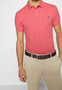 Rotes Polohemd mit Kragen und kurzen Ärmeln, mit einem kleinen blauen Logo. Getragen mit beigen Hosen und einem dunkelbraunen Gürtel.