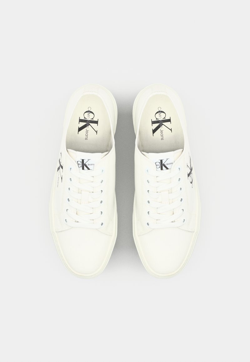 Baskets en toile blanches avec des bouts ronds, des lacets plats et des accents de logo. Texture lisse avec un design minimaliste et des détails de couture subtils.