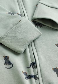 Vêtement zippé vert clair en tissu doux, présentant un motif de léopard répétitif en bleu foncé et des accents orange. Les poignets sont côtelés.
