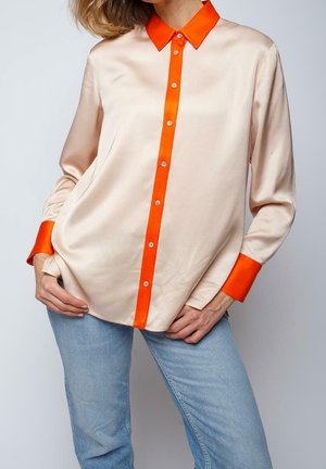 Kvinde iført en cremefarvet satinbluse med lyseorange krave, knapstolpe og manchetter kombineret med lyseblå jeans.