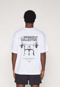 Άσπρο βαμβακερό μπλουζάκι με μεγάλο γραφικό τύπωμα πάγκου άρσης βαρών και μπάρας στη ράχη. Το κείμενο αναγράφει «The WORKOUT COLLECTIVE.»