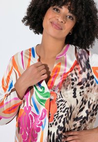 Mujer con cabello rizado, vistiendo una blusa colorida con patrones florales y de animales, tocando suavemente el cuello, sonriendo suavemente.