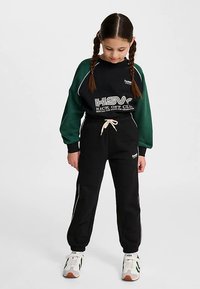 Czarny i zielony zestaw sportowy składający się z bluzy z okrągłym dekoltem z logo i napisem, zestawionej z czarnymi dresami z bocznymi paskami. Białe trampki.