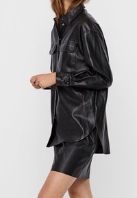 Chemise en simili cuir noir, semi-structurée avec deux poches poitrine, un col et des manches longues, associée à une jupe assortie.