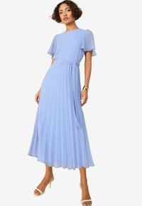 Robe plissée bleu clair avec des manches courtes et flottantes, une taille cintrée et une ceinture nouée. Fabriquée en tissu fluide, longueur cheville avec un léger éclat.