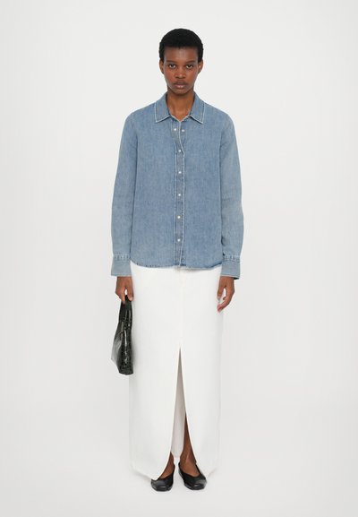 Light blue denim shirt with a collar, button front, and long sleeves, paired with a white maxi skirt featuring a front slit. Black shoes and a black bag.