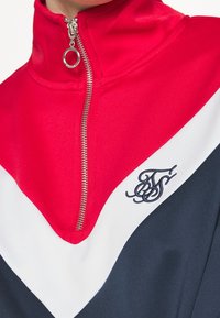 Pullover zip con collo alto in rosso e blu navy, con strisce diagonali bianche e logo ricamato. Tessuto liscio e accessori lucidi.