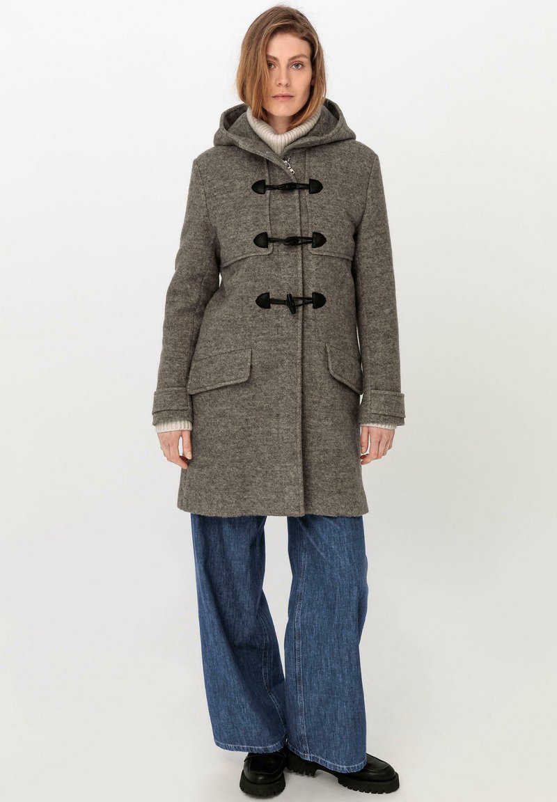 Grauester Wollmantel mit Kapuze, Toggle-Verschlüssen und zwei Fronttaschen. Kombiniert mit weiter geschnittenen Denimhosen und schwarzen klobigen Schuhen.