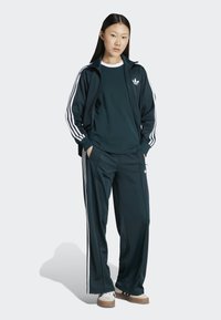 Dark green athletic tracksuit with white stripes on sleeves and pants. Featuring a zip-up jacket, long sleeves, and relaxed-fit trousers.