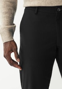 Pantalon noir sur mesure à la texture lisse, doté d'une fermeture standard à bouton et d'une poche latérale, accompagné d'un pull beige clair.