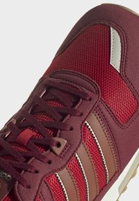 Tmavě červené a červené tenisky s hnědými koženými pruhy, látkovou síťovinou a tmavě červenými tkaničkami na bílém pozadí.