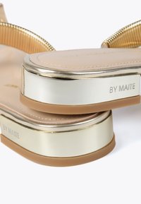 Beige-sandaalit, joissa on metalliset kultaiset yksityiskohdat ja sileä pinta. Sisältää paksun korokkeen ja merkinnän "BY MAITE" sivulla.