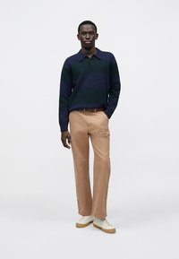 Πλεκτό πόλο μπλουζάκι με λευκές και πράσινες ρίγες και γιακά, συνδυασμένο με ανοιχτό καφέ chino παντελόνι και λευκά αθλητικά παπούτσια με μπεζ λεπτομέρειες.