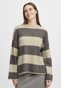 Pull rayé en beige et gris foncé, fabriqué à partir d'un matériau tricoté doux. Il présente un col rond et des manches larges et amples. Fini texturé.