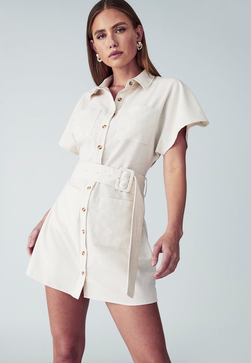 Krótką sukienkę z paskiem z jasnego beżowego materiału, z przodem zapinanym na guziki, dwiema kieszeniami na piersi oraz lekko rozkloszowanym fasonem spódnicy.