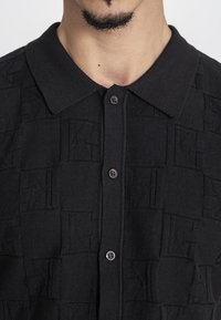 Polo nero con un motivo testurizzato di intersezioni di design con la lettera "K", colletto e chiusura frontale con bottoni scuri.