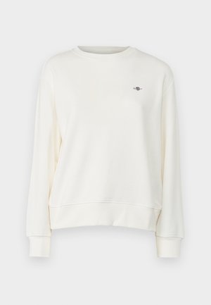 Sudadera de cuello redondo en color crema, con mangas largas y un sutil acento de logo en el pecho. Textura de tela suave y lisa.